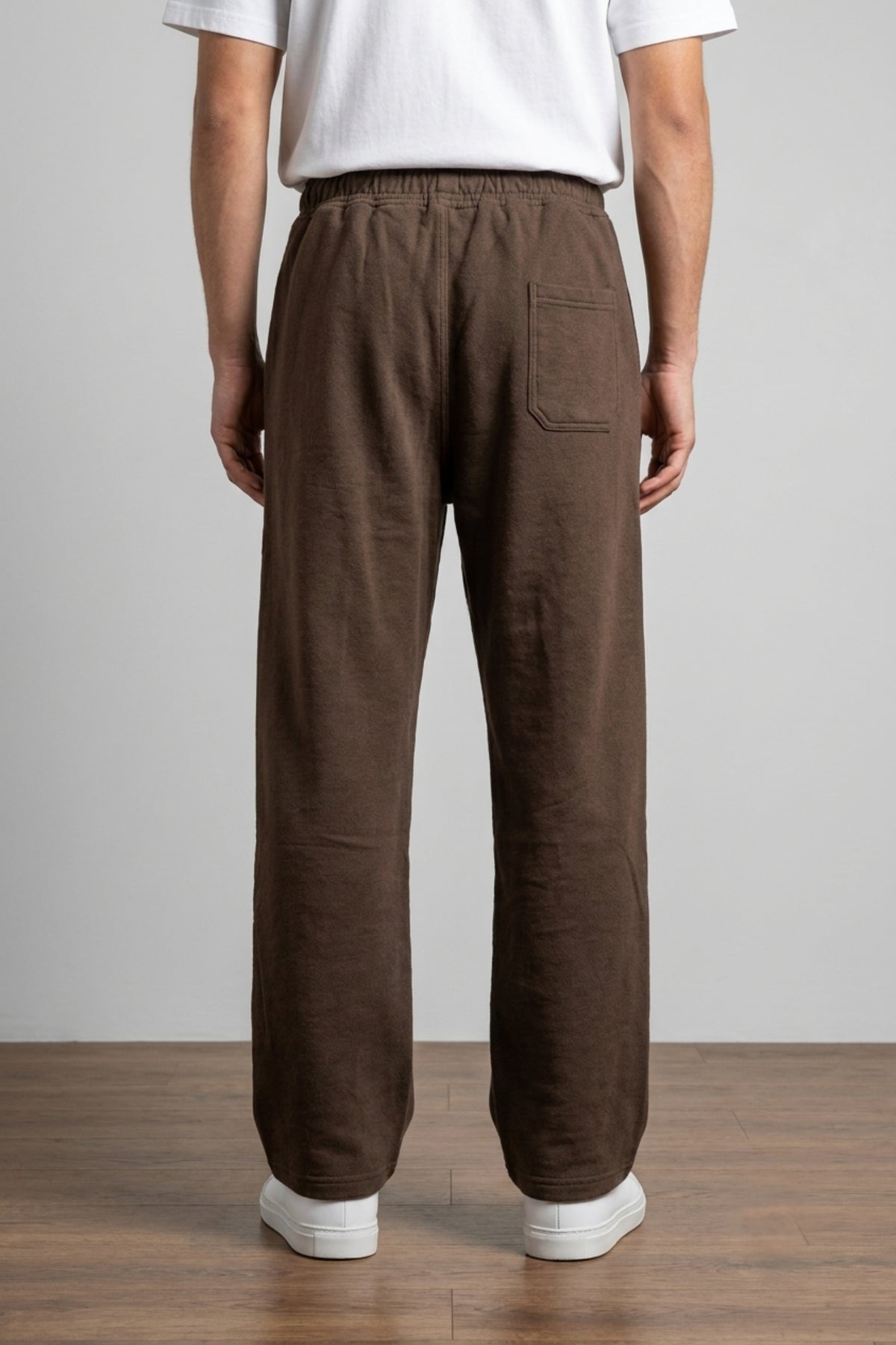 Rear view of a man wearing dark brown baggy terry trousers with a functional back pocket, relaxed fit, and white minimalist sneakers.